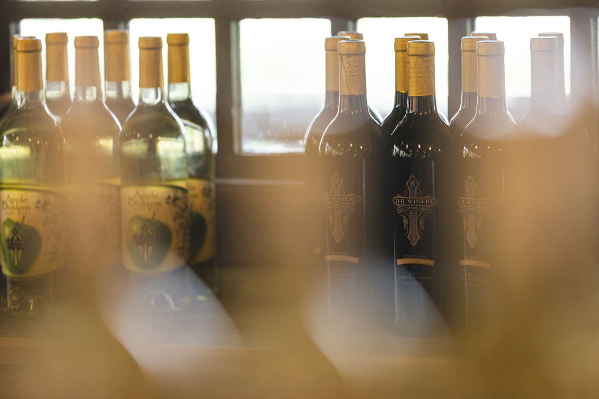 Wine bottles sit on a shelf at the Winery at Holy Cross Abbey behind a slightly blurry object. Behind the bottles are windows letting in natural light.