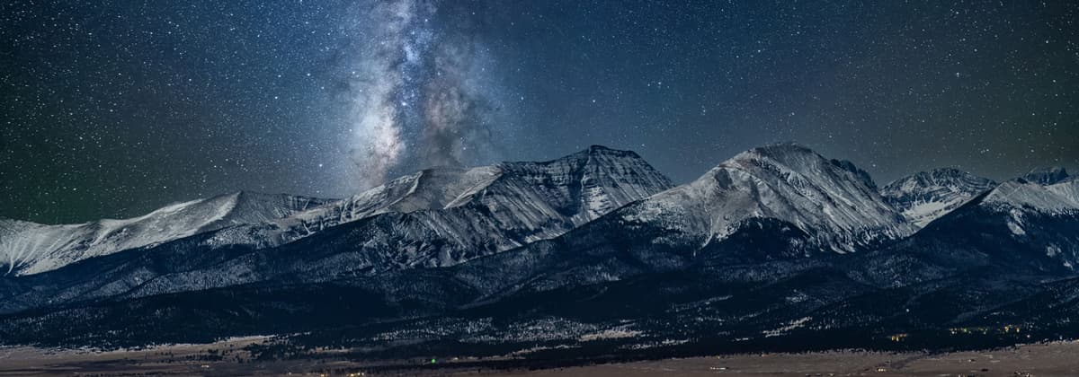 A panoramic photo of the snowcapped Sangre de Cristo Mountains in Westcliffe, Colorado, at night. The sky above the mountains is dark gray and black with what looks like thousands of little glowing stars speckling the sky. Slightly to the left of center of the photo is a vertical cluster of multi-colored stars making up the beautiful Milky Way Galaxy.