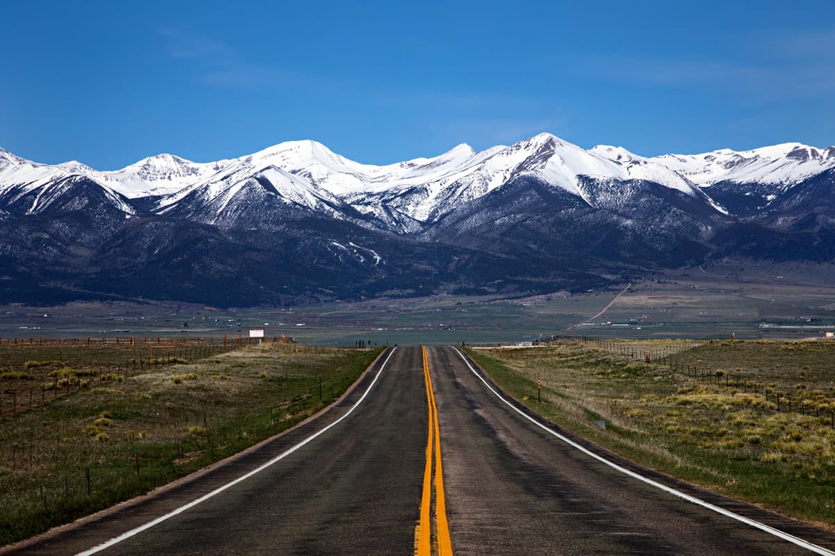 In the foreground of the photo is a black road with white lanes and yellow lanes stretching to the middle of the photo, where the road drops off. Beside the road on either side are fields of green grass. In the background of the photo is the large, deep-blue and snowcapped Sangre de Cristo Mountains of the Frontier Pathways scenic byway in Westcliffe, Colorado, looming majestically over the town under dusty-blue skies.