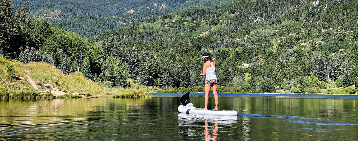 A standup paddleboarder dressed in summer clothing stands on a paddleboard on Lake Isabel in Westcliffe, Colorado. The waters gently ripple under the water craft and in the background are hills upon hills of pine trees and grass.