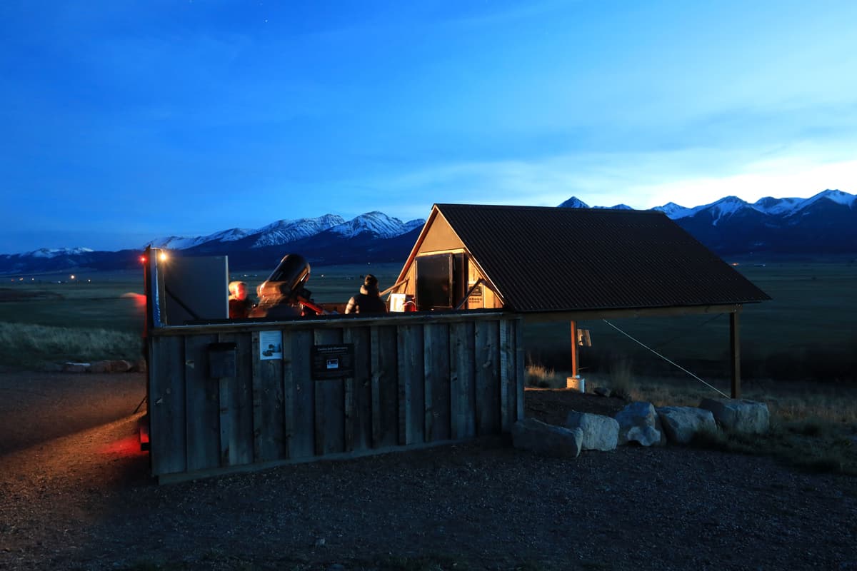 The Smokey Jack Observatory, a small roofed structure with wood-paneled walls, stands on a patch of dirt in Westcliffe, Colorado. The sky above the observatory is deep blue after a recent sunset. The Sangre de Cristo Mountains are snowcapped and majestic in the background of the photo.