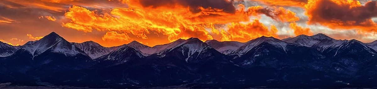 A striking panoramic photo of the Sangre de Cristo Mountains, snowcapped and dark blue. Above the mountains are orange, pink and yellow whispy clouds, looking like licks of fire above the mountains during sunset in Westcliffe, Colorado.