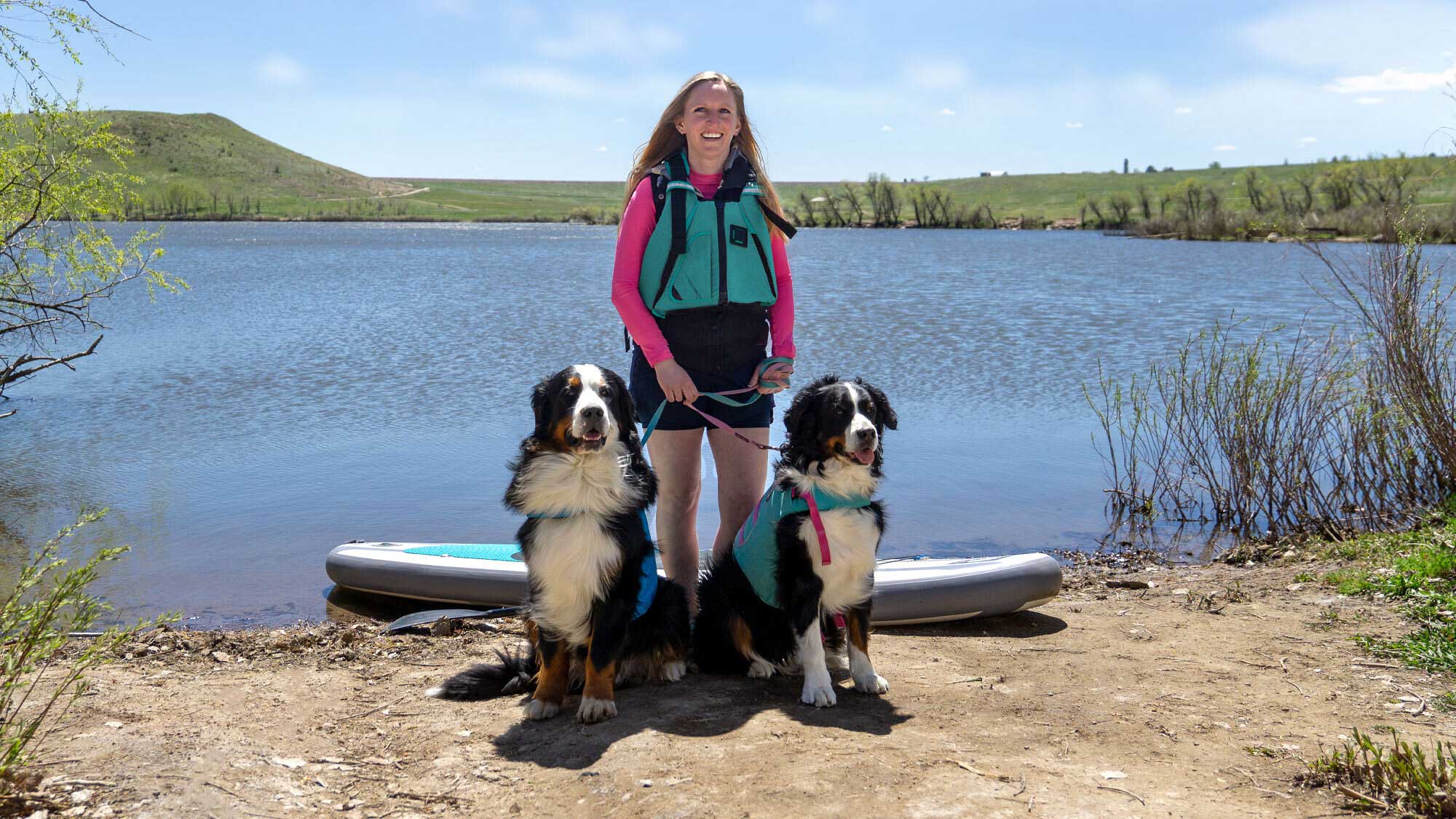 A woman and two dogs, all three in life vests, stand by their standip paddleboard on the shore of a lake