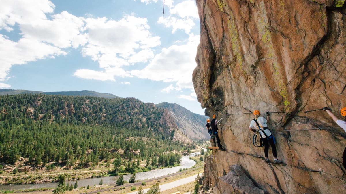 Four people in protective gear who are tethered shuffle across a large cliff face dotted with moss on the Colorado Via Ferrata course with Arkansas Valley Adventures Rafting & Zipline