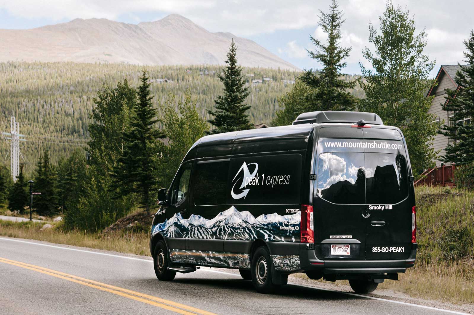 A black sprinter van with the Peak 1 Express logo drives a road toward a mountain