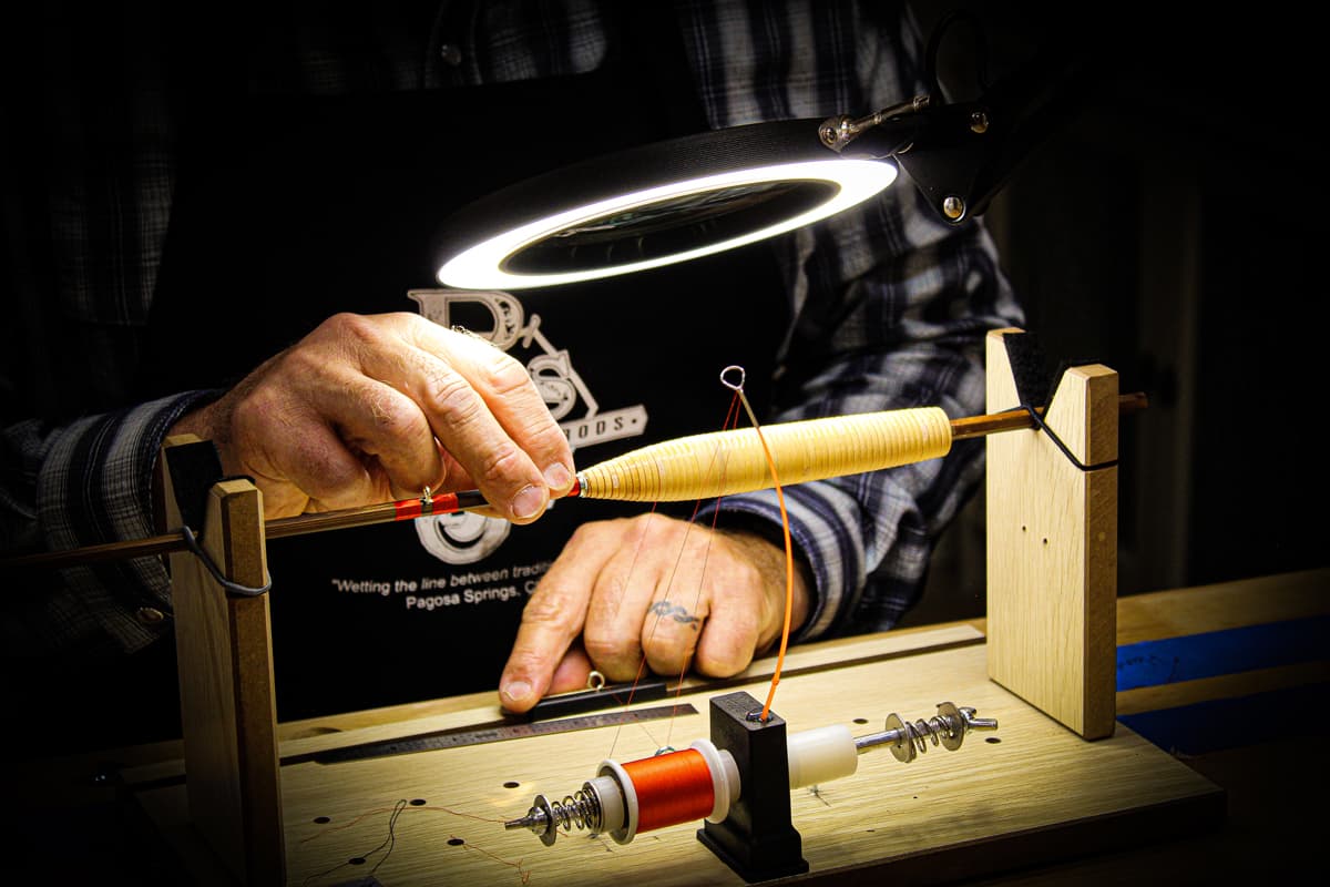 A large, circular light blasts light onto pieces of equipment for a person building a handcrafted fly-fishing rod with PJ's Fine Bamboo Rods in Pagosa Springs. They are meticulously threading super-fine rattan thread into the handle of the fly-fishing rod.