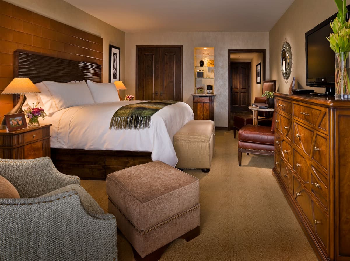 A rustic and warmly decorated luxurious room in Madeline Hotel & Residences in Telluride. The bed is large and adorned in white linen, sat against a dark-wood bedframe, flush against more warm-brown walls. There are wooden and brown decorations around the room, including a minibar, a few chairs, a dresser, and a nightstand. The lamp on the nightstand bathes the side of the bed in a warm golden glow.
