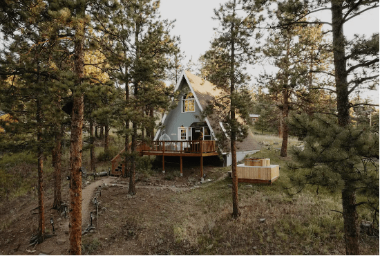A mint-green Aframe in Bailey, Colorado, stands among pine trees with an open clearing in front of it.