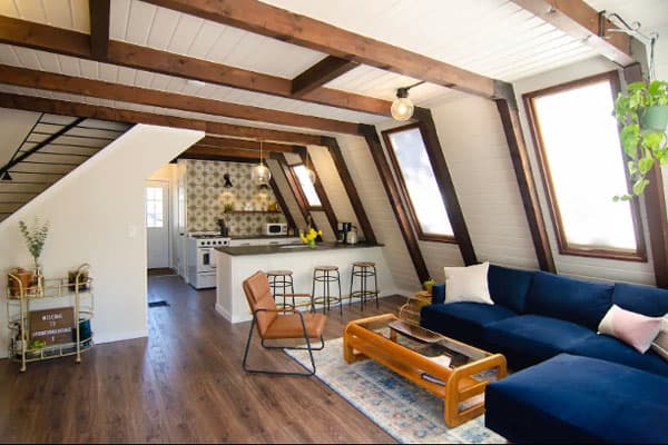 The interior of the Bailey A-Frame. The roof is in a geometrical shape, white with accents of dark brown wood. There is a long L-shaped dark blue couch with light pink pillows and a wooden coffee table with a brown chair next to it. In the background is a small kitchen with patterned wallpaper, several appliances, and three wire-legged stools at the bar. To the left is a golden metal bar cart with fake plants, alcohol bottles, and a welcome message on a little board.
