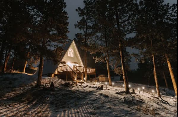 A nighttime photo of the exterior of the Bailey A-Frame. There are twinkling lights along the path winding up to the front door of the a-frame, which is lit in the glow of the porch light. There is light snow on the ground and sparse, tall pine trees.