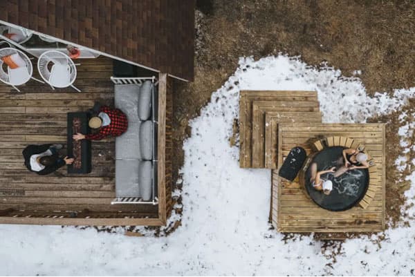 An aerial shot of the cedar hot tub and half of the patio at the Bailey A-Frame. There are two people in the hot tub and two people tending to the patio firepit on the patio. The patio is decorated with a few white wire chairs and a large gray couch. The brown ground is covered mostly in snow.