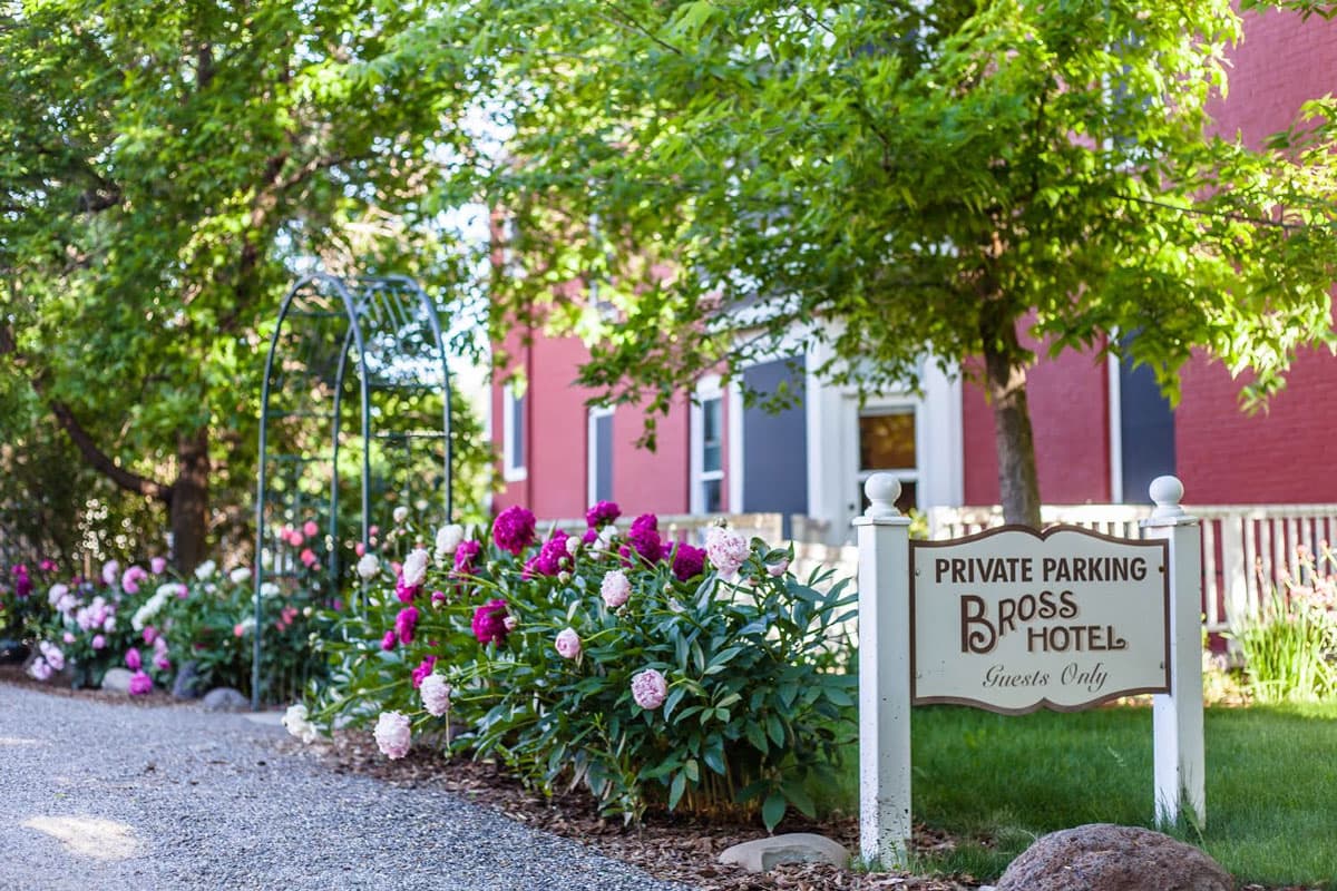 The crimson Bross Hotel Bed & Breakfast is in the background, with a sign, verdant tree leaves and blooming peonies in shades of pink.