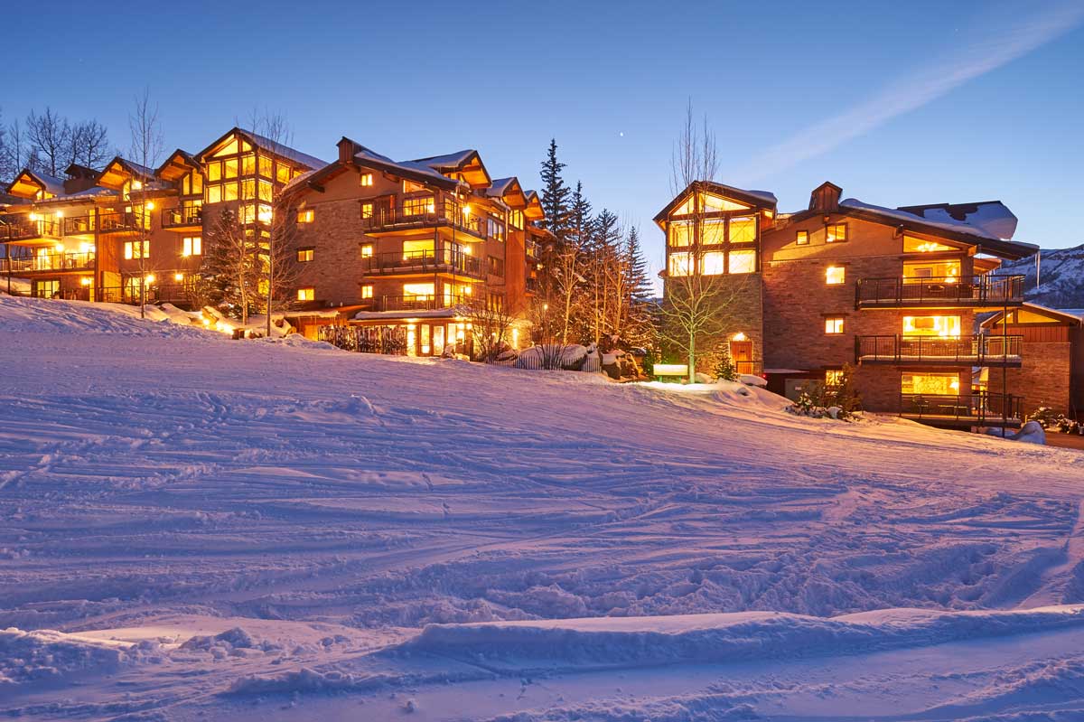 Night is setting in at Snowmass Village and lights of The Crestwood Condominiums glow bright. The sky above is clear and there is snow on the ground.