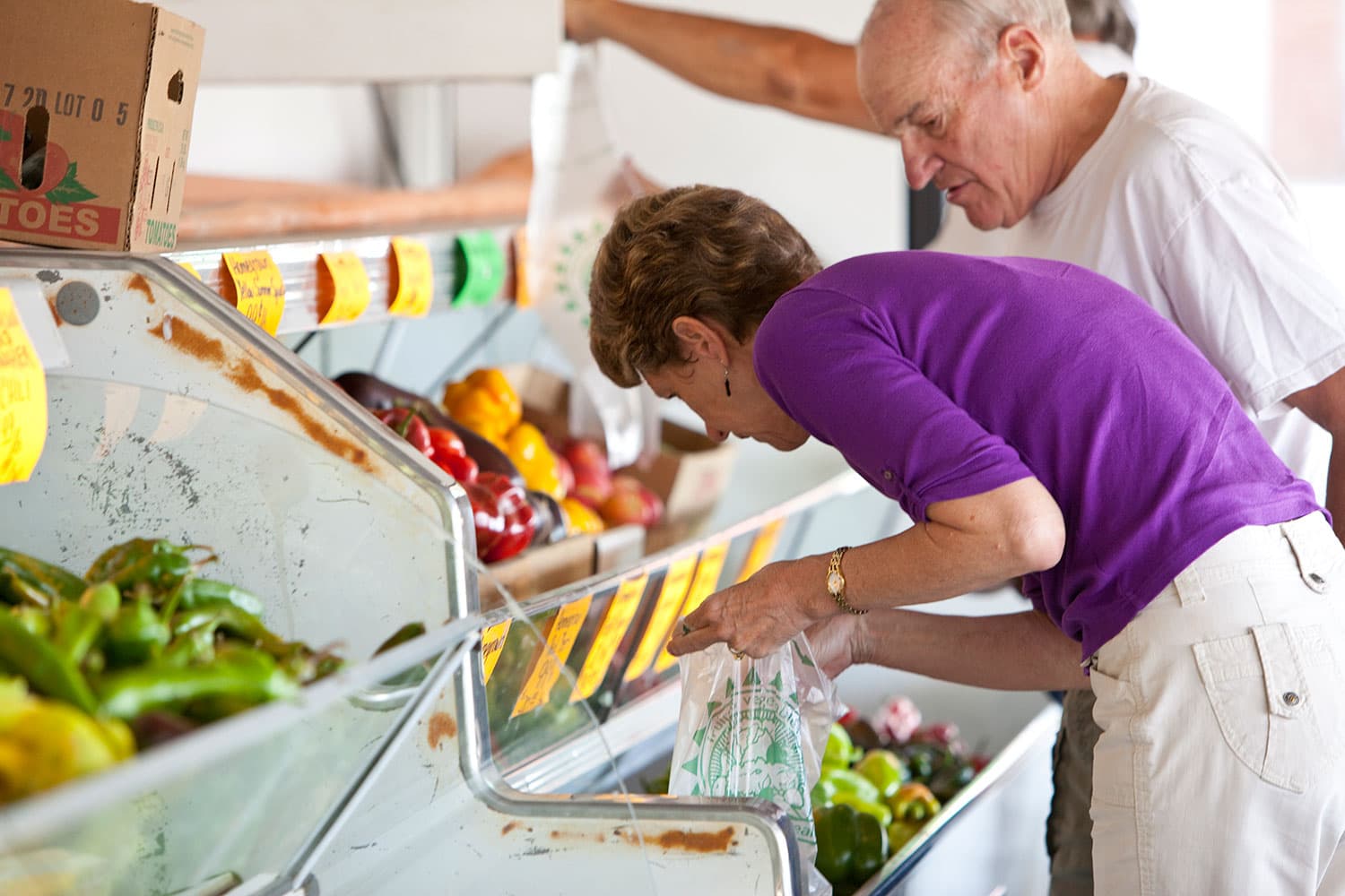 A woman in a bright-purple shirt with khaki pants and a man in a white-shirt look into a case of fresh produce.