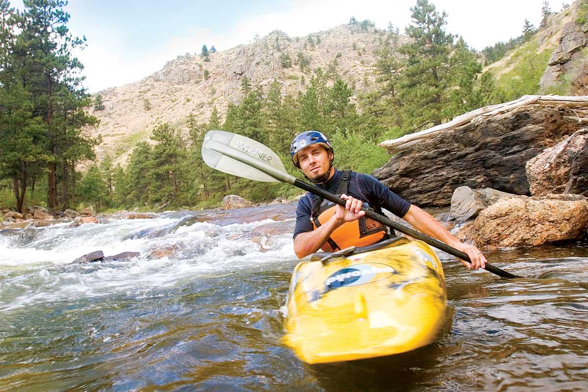 A yellow kayaker paddles along the Cache la Poudre River, dodging rocks and rapids; he wears a helmet