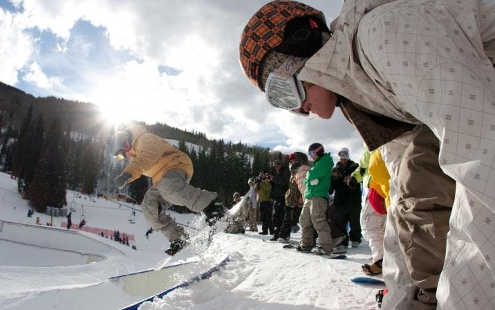 A gathering of snowboarders in helmets and googles waits their turn to hit the halfpipe as the sun sets behind the mountain