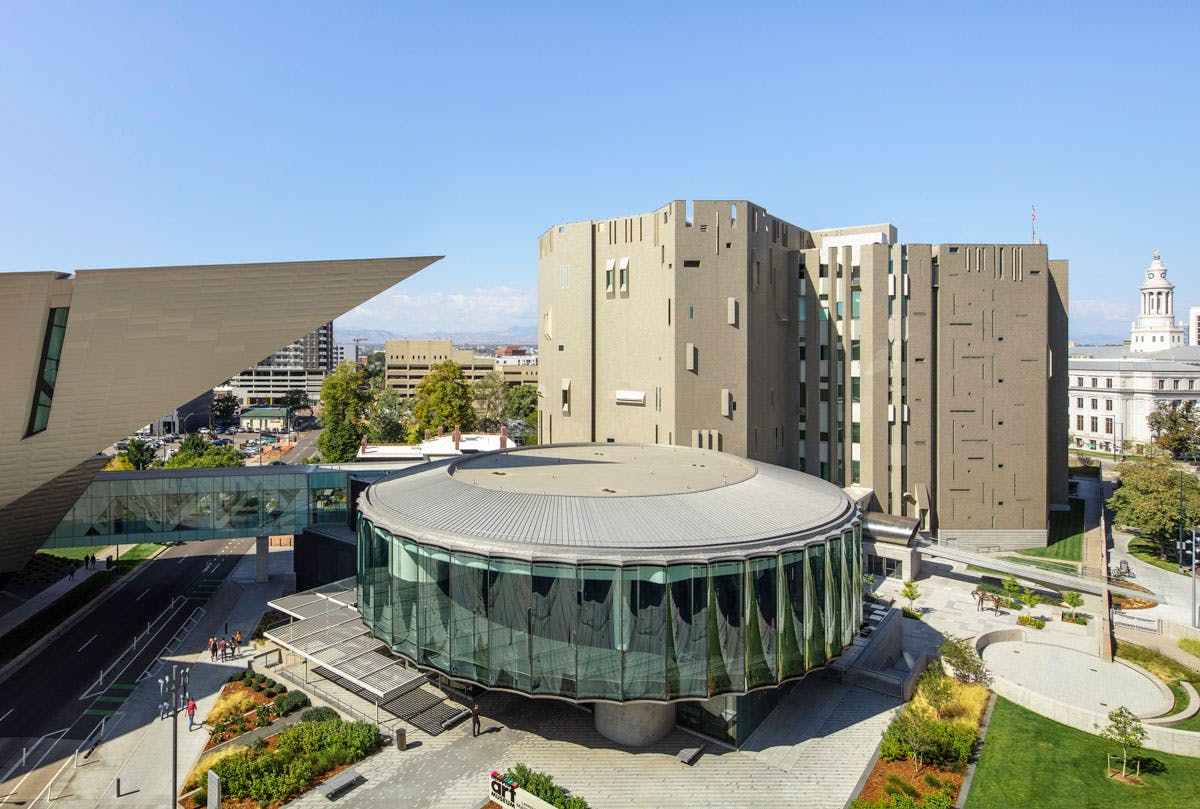 A view of the Denver Art Museum's three buildings on a light-blue skied summer's day.  The triangular point of the main building sits above a round glass building with another geometric five-story building behind it.