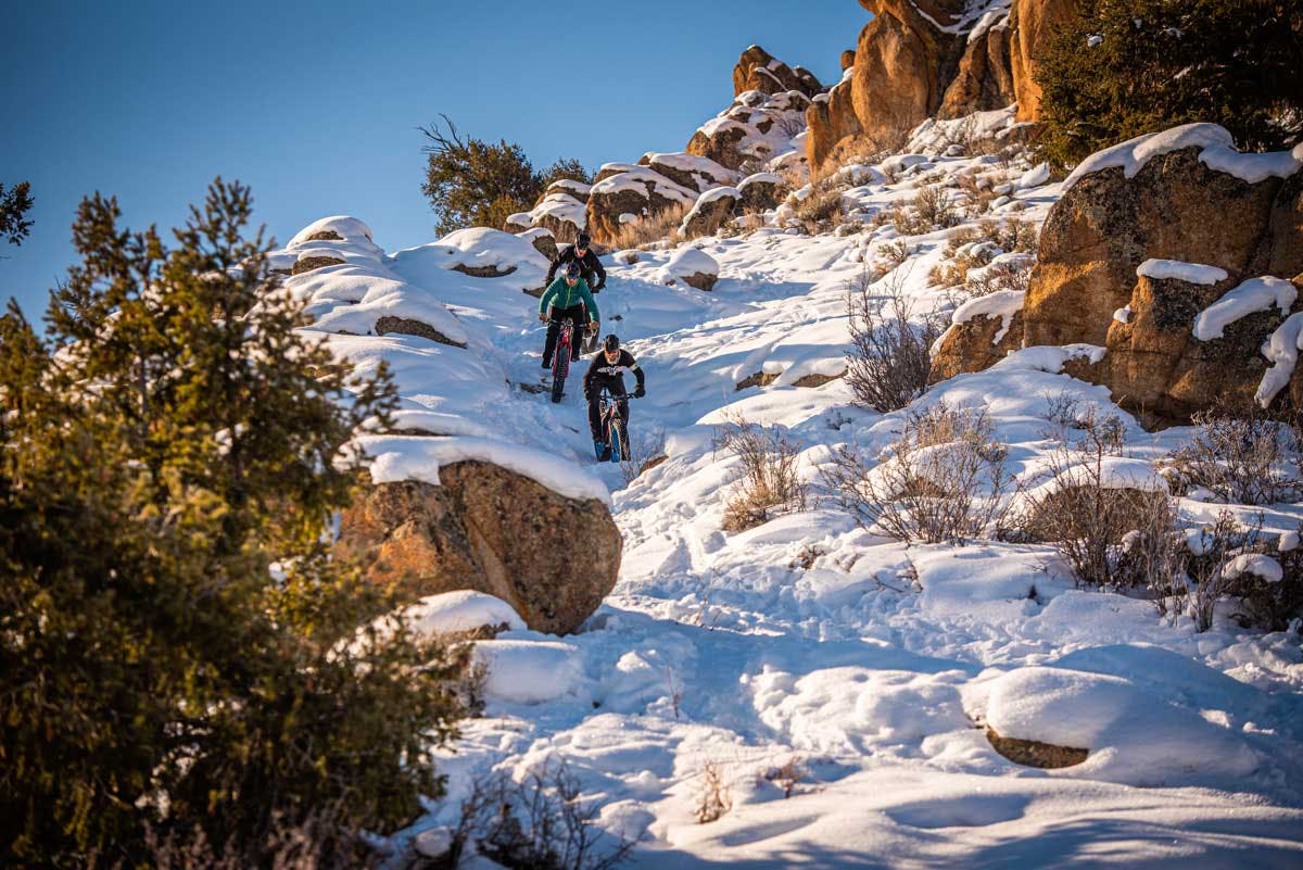 Three fat bikers pedal downhill down a snow-covered path in winter. There are red rocks dusted with snow and there's bushes and trees sticking out of the frozen earth.