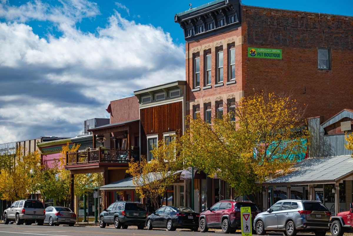 Against the backdrop of a blue sky with white clouds, the colorful storefronts of downtown Gunnison sit in the early fall with golden-leafed trees.