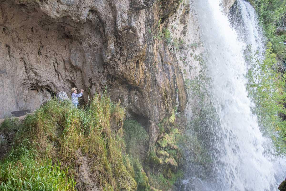 A hiker stands on a flat patch of land and takes a photo of a cascading waterfall at Rifle Falls State Park in Colorado.