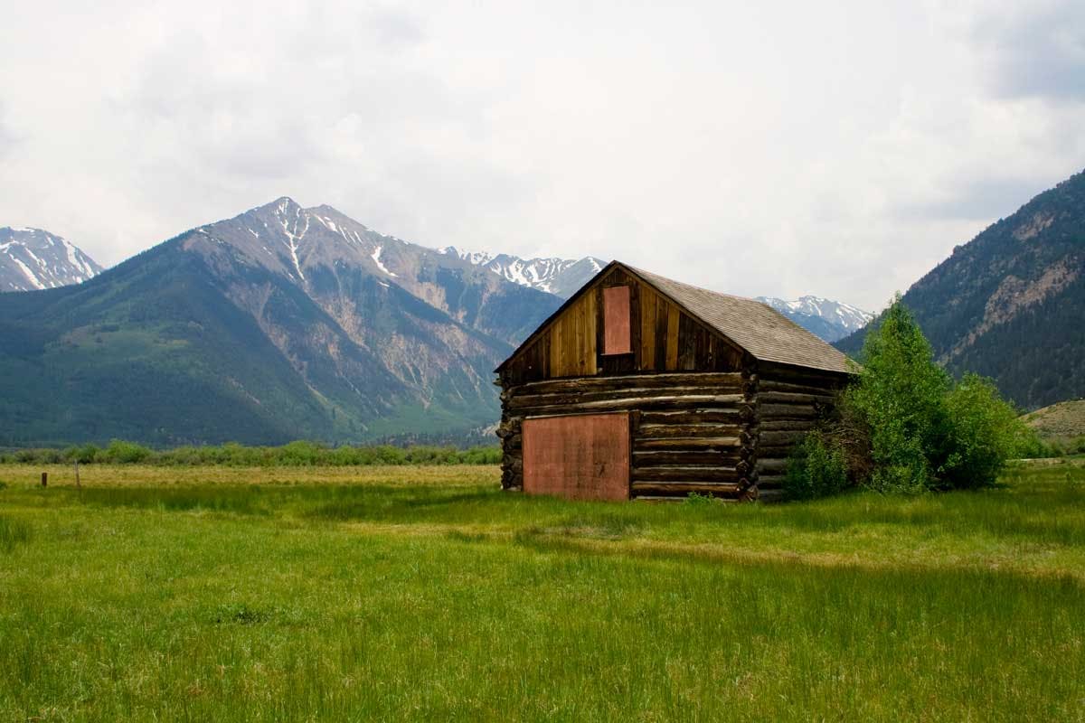 An old log cabin stands alone in a field near Twin Lakes. Behind it are tall mountains with the remains of winter snow.