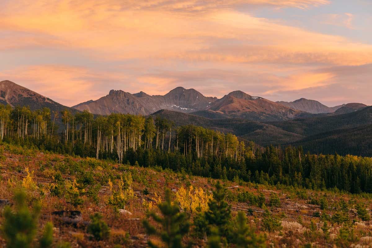 The clouds and sky at State Forest State Park are tinged with pinks, dusky purples and light yellows as the sun sets. Mountains and pine trees below stand tall.