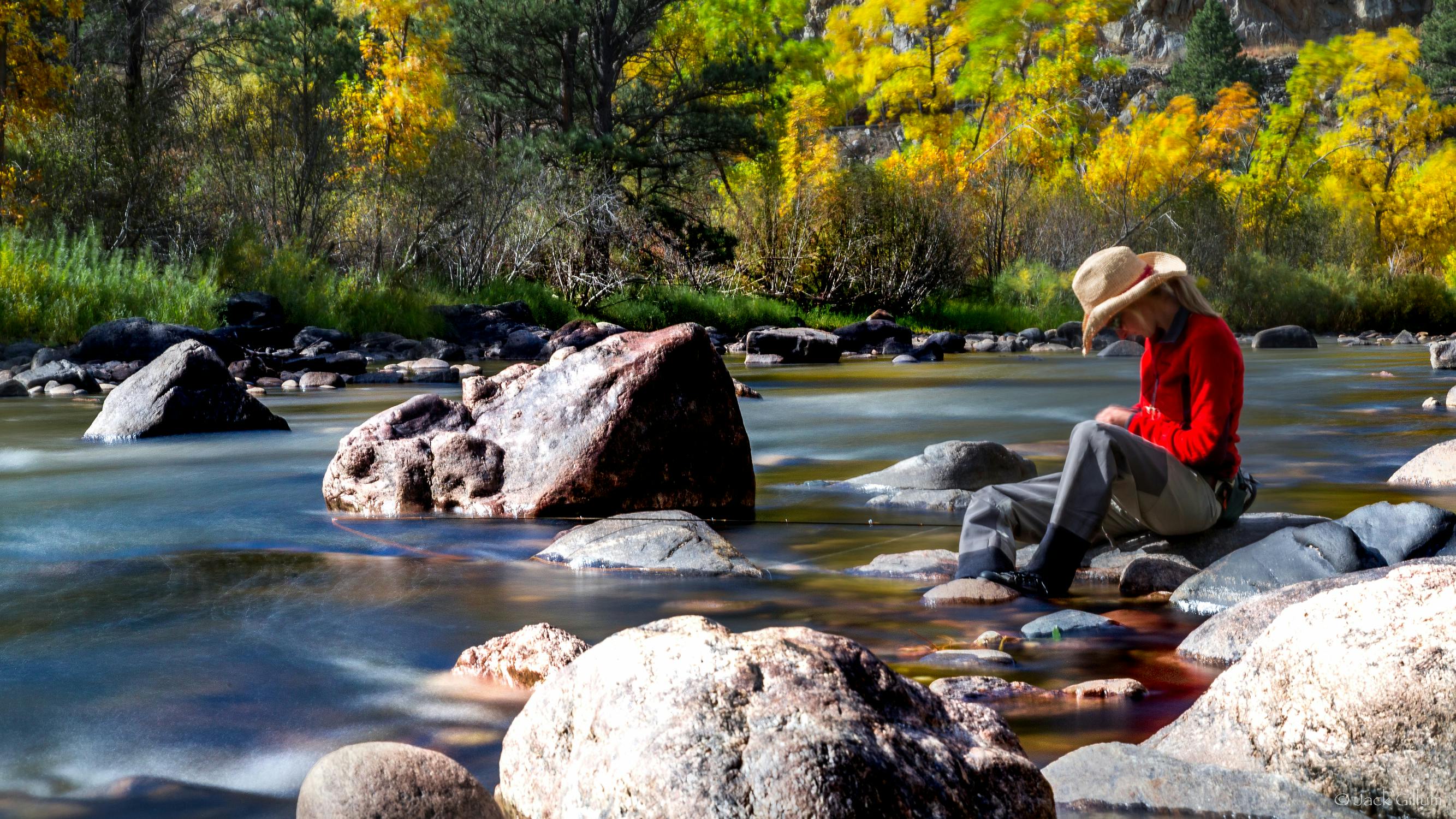 A fisher wears waders, a red sweater and a wide-brim hat and sits on a rock near the shore of the Cache la Poudre River.