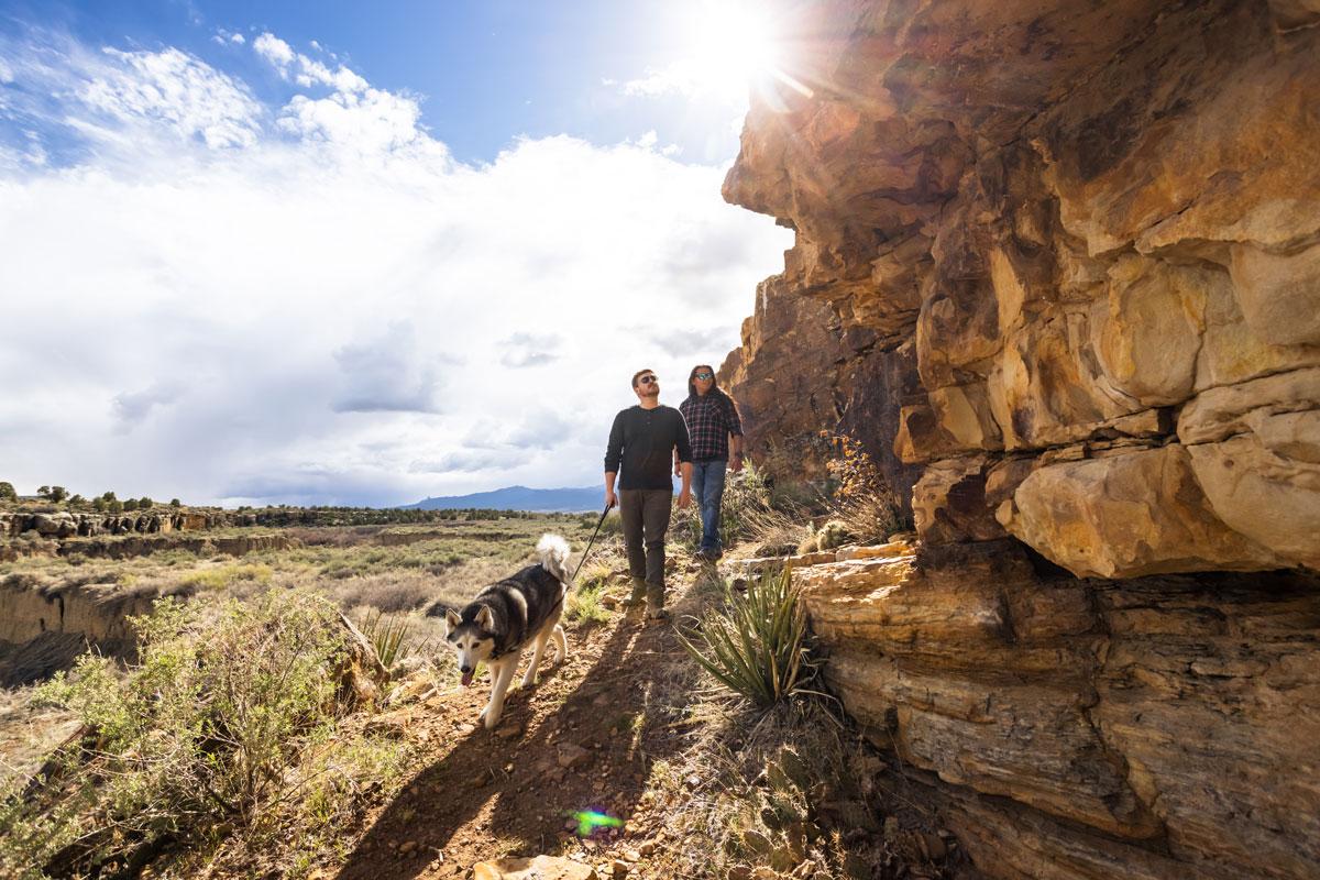 Two people hike and walk a dog along a rocky path. To their right, a wide open valley with a blue sky and lots of white clouds. To their right a rocky face with multiple levels.