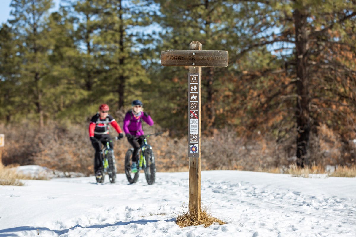 Two fat bikers cross a snowy path. In the foreground a wooden t-shaped sign with multiple markings.