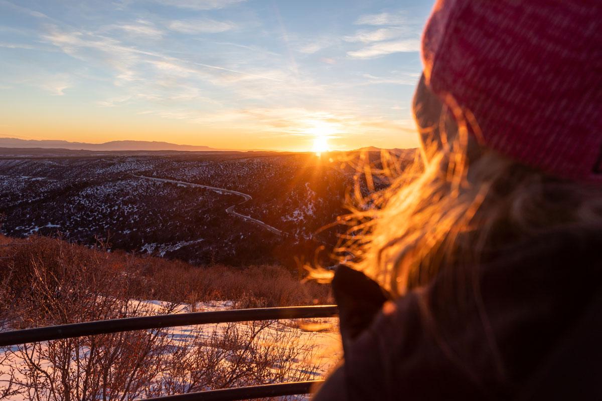 A person's red beanie, hair and shoulder in a jacket are shown at an observation point with a metal rail. The person looks out at the sunset over Cortez. There is a winding road in the distance and the ground is covered in snow. 