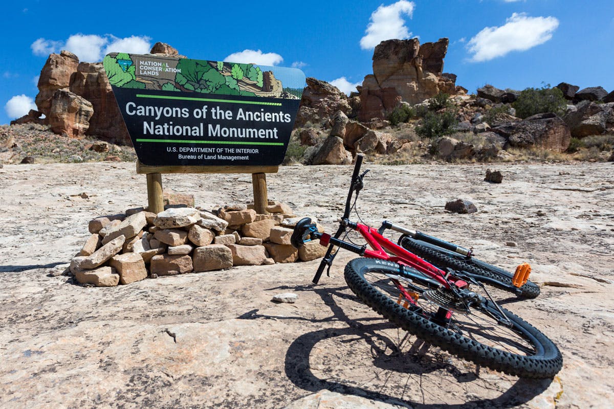 A rocky area with a red bike on its side. In the background rock formations sit and in the middle a sign reads "Canyons of the Ancients National Monument" and beneath that it says "U.S. Department of the Interior Bureau of Land Management."