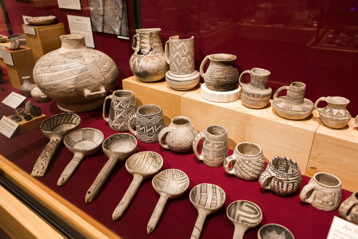 A museum display, behind glass with a red wall and red bottom with three rows of pottery artifacts. 