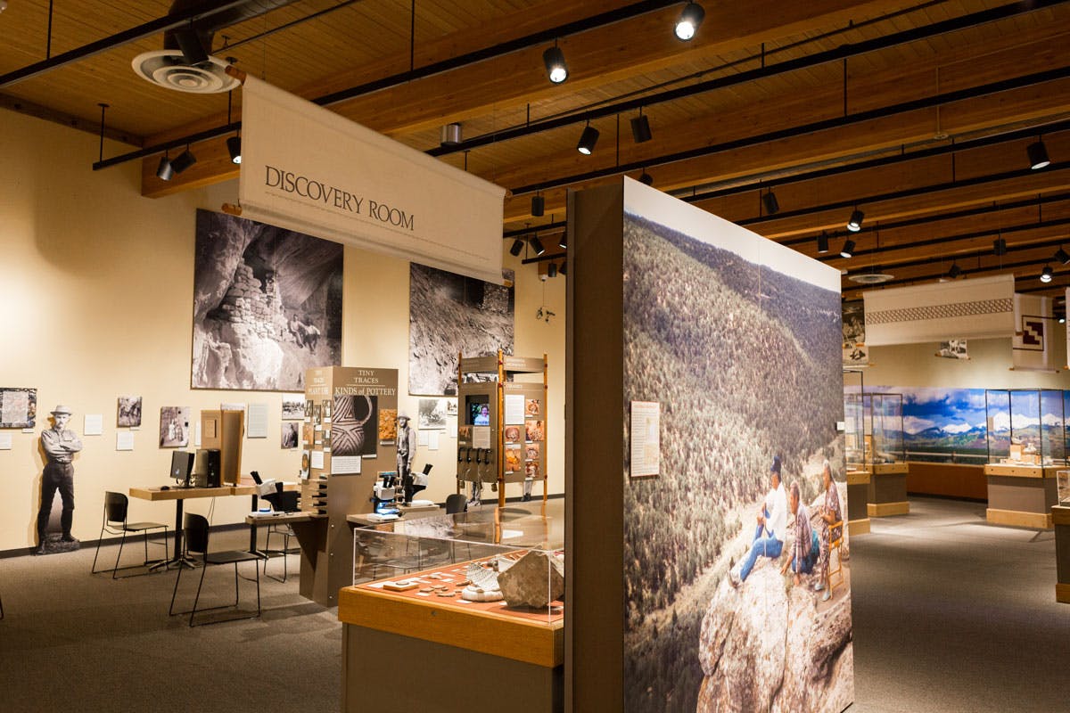 A museum room with a sign that says "Discover Room" hanging from the ceiling. The room is a museum gallery with images, glass displays and tables with computers.