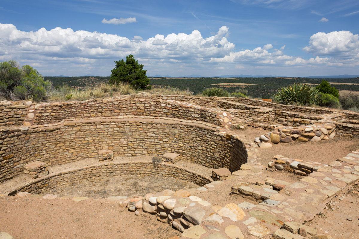 A stone, man-made hole in the ground is surrounded by small stone walls, green scrubby vegetation and a blue sky with white clouds at a Canyons of the Ancients National Monument site.