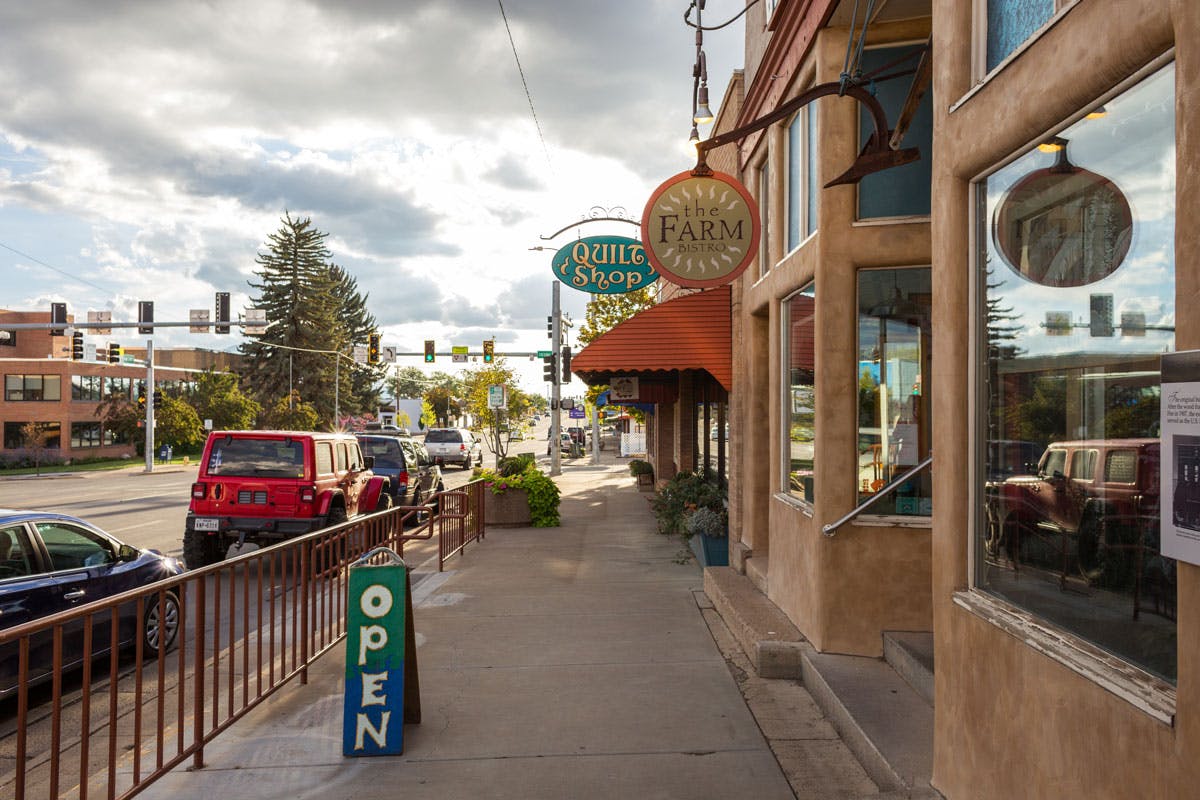 A street in downtown Cortez with an a-frame sign that is vertical painted half green and half blue that says "Open" on the sidewalk. On the right two businesses, one with a sign that reads "The Farm" and the other says "Quilt Shop." Their are cars parallel parked on the left with evergreen trees and brick buildings in the distance.