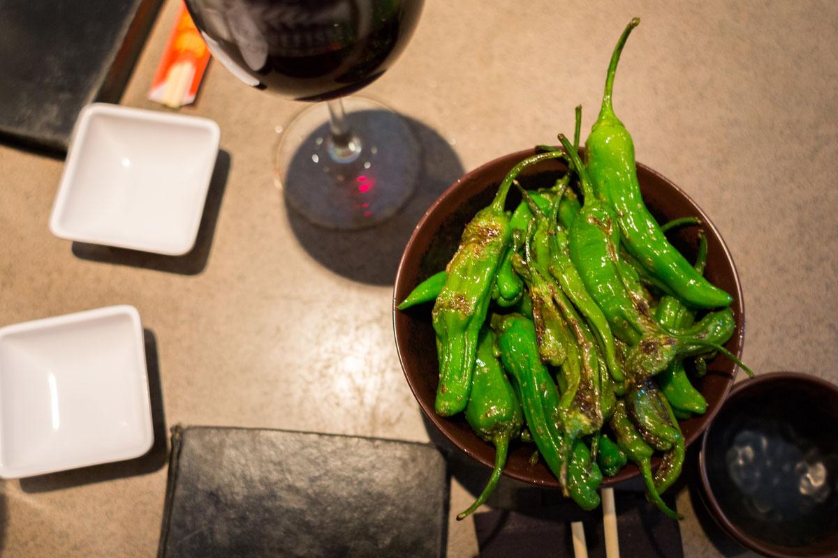 Bright green, grilled peppers in a bowl on a table. There's a partial wine glass and two square white dishes meant for soy sauce at Stonefish Sushi & More.