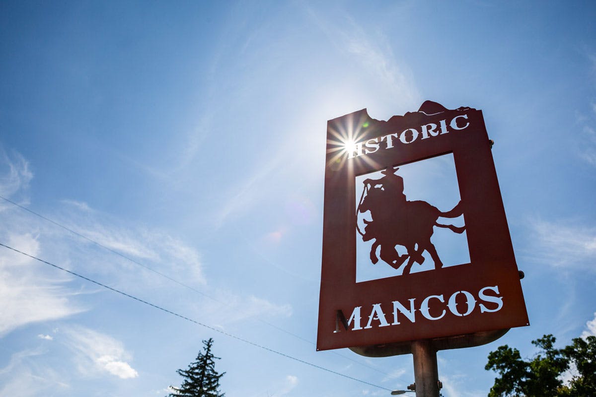 A metal cutout sign with a person wearing a cowboy hat on a horse says "Historic Mancos" in the blue sky.