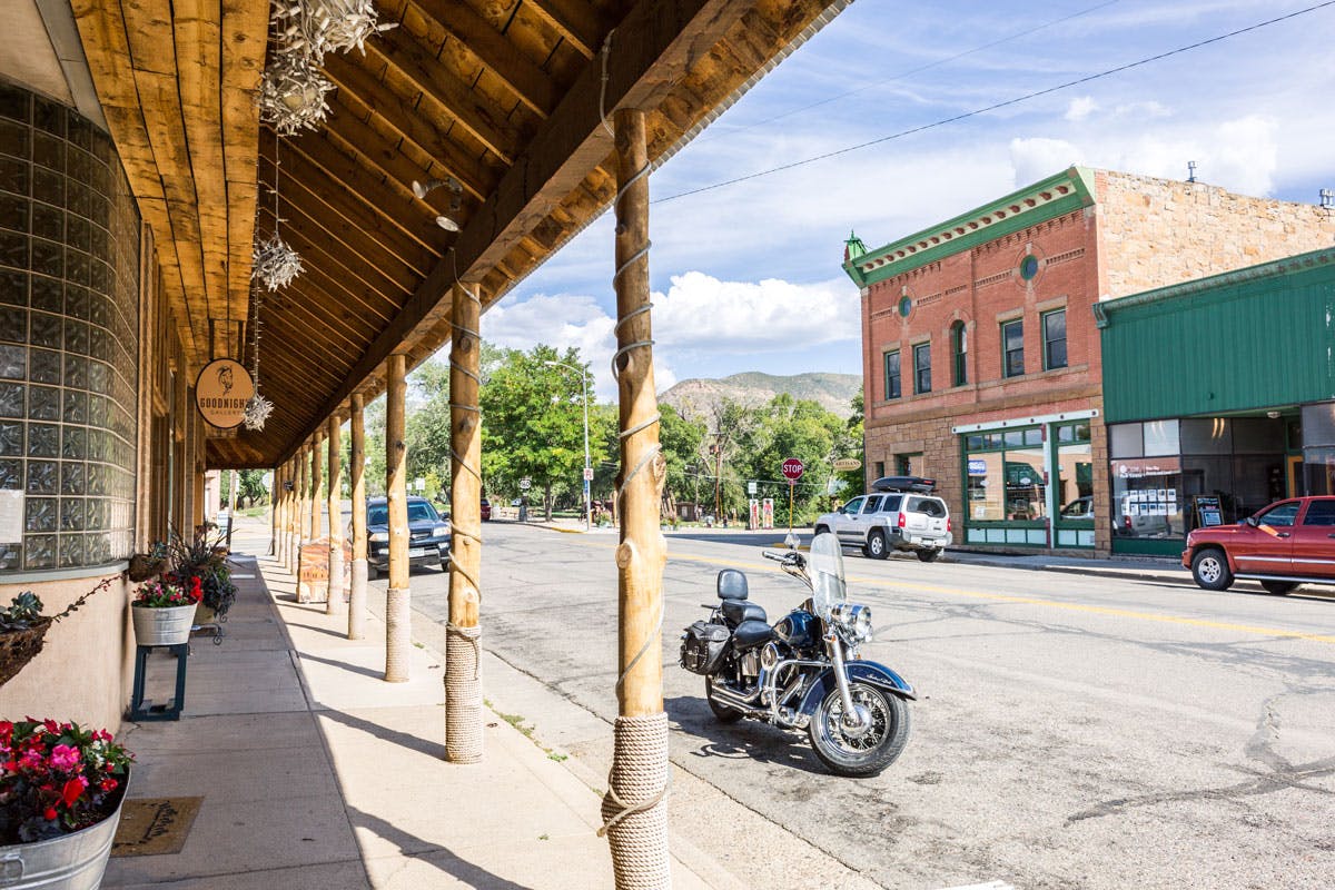 A motorcycle is parked out front of a storefront with glass block windows and a wooden overhang with columns. Across the street, a two-story red-brick Western-style building sits with cars in front of it. In the distance mountains can be seen with green trees and blue skies.