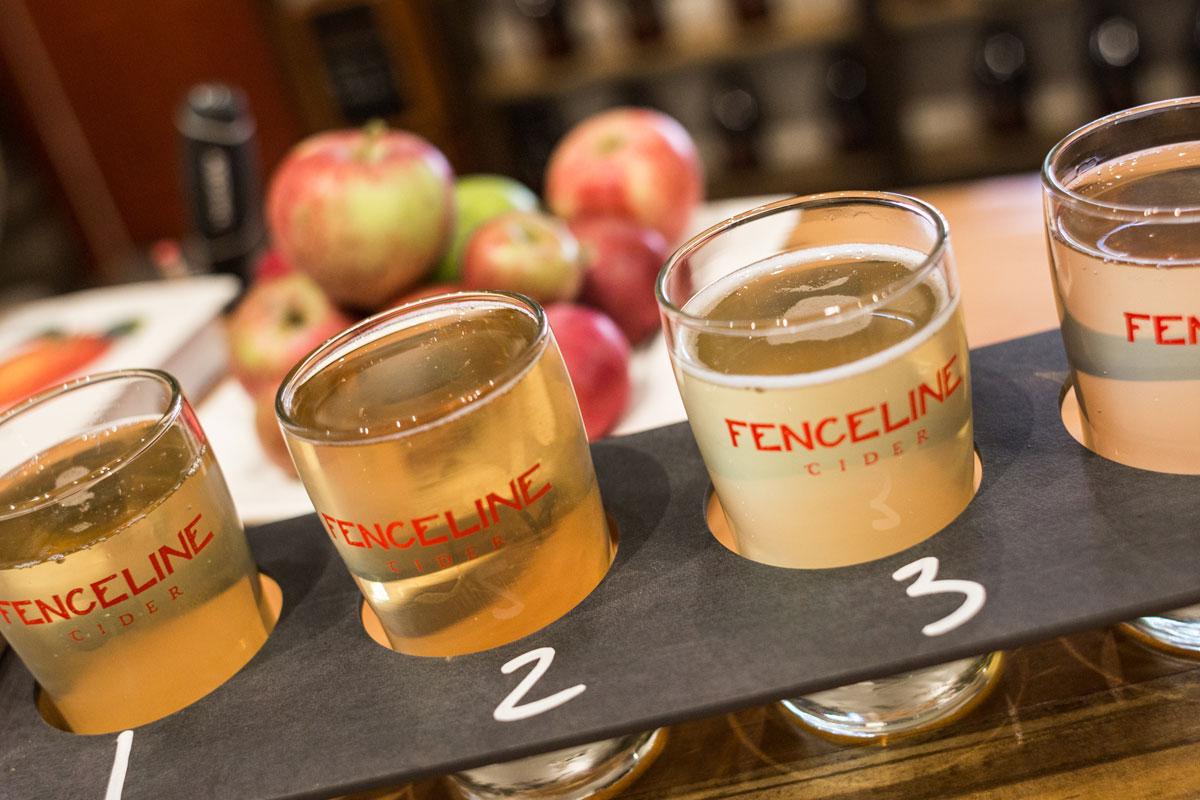 A flight with glasses of different Fenceline Ciders sit in a tray that are numbered. In the background a pile of apples.