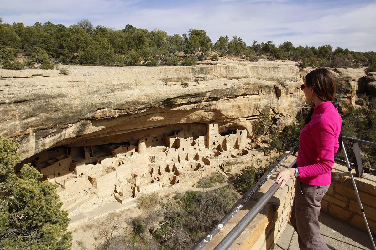 A person in a hot=pink jacket and brown pants hold onto a metal rail while overlooking sand-colored cliff dwellings at Mesa Verde National Park.