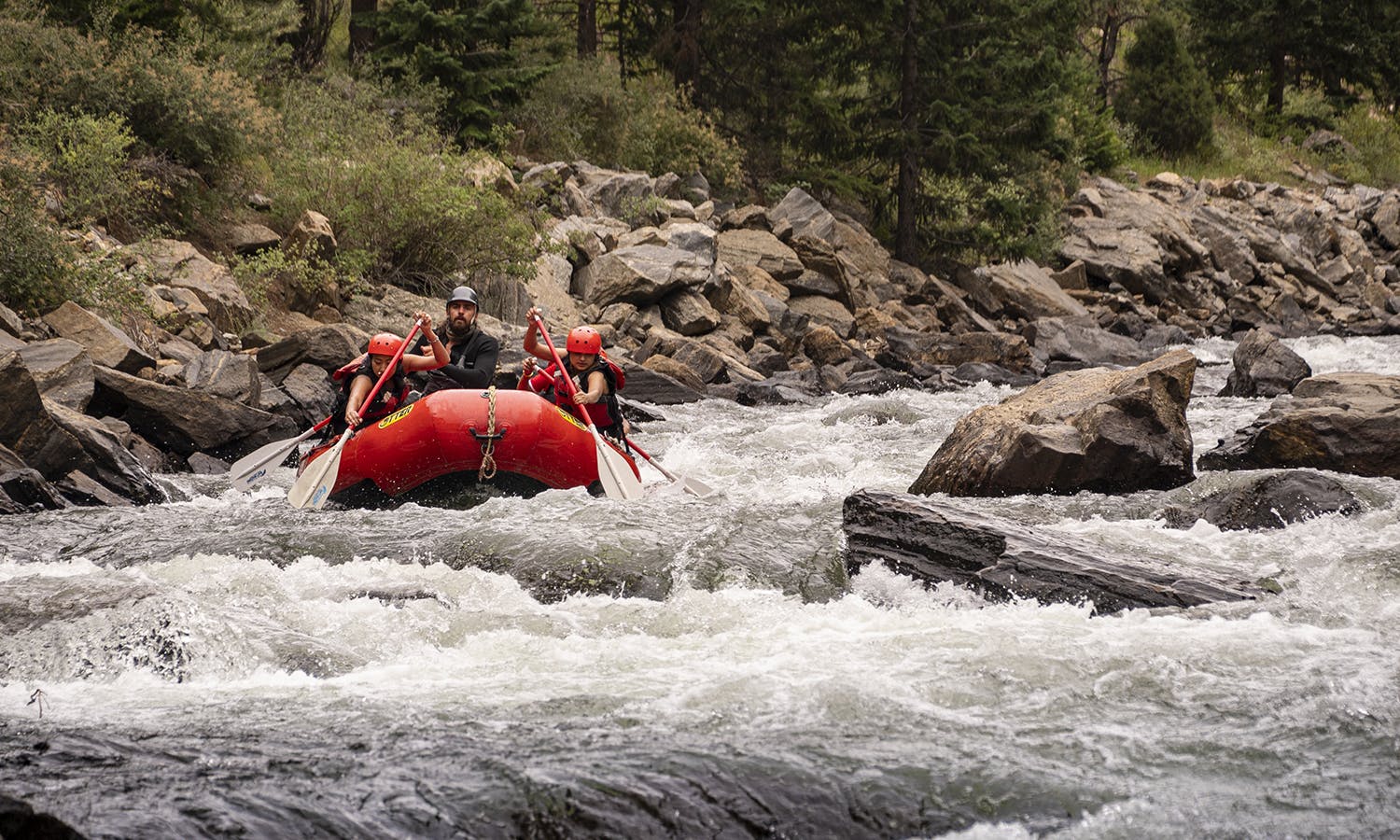 Red inflatable raft with three people whitewater rafting through rapids.