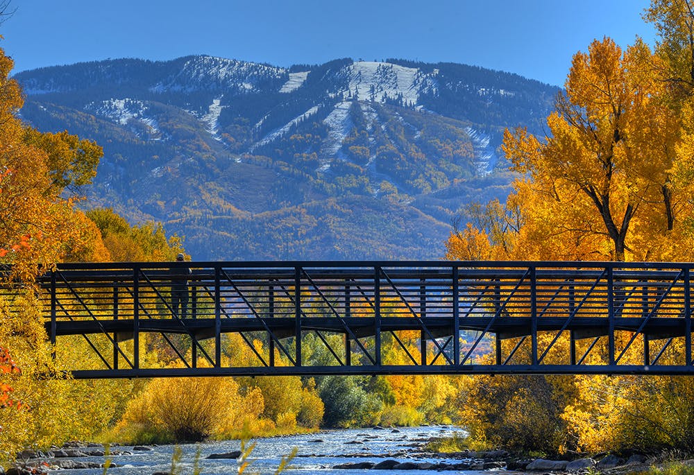 Yampa River running through the heart of Steamboat with the mountains in the background. A pedestrian bridge spans the river and there are trees with golden fall leaves on either bank. 