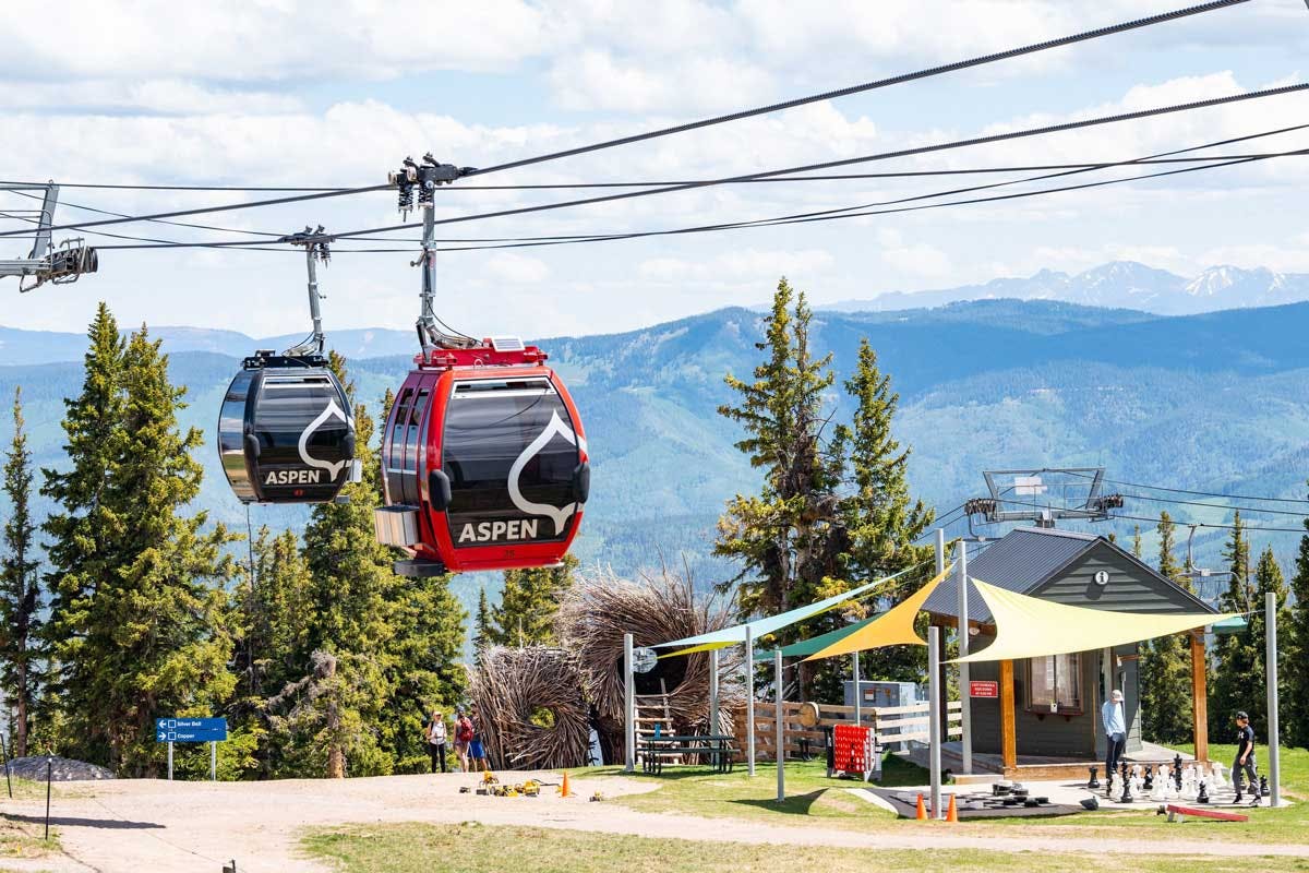 Two gondolas, one red and one black, both with the Aspen logo on them, carry passengeres above a tented picnic area. Green and blue mountains are in the background.