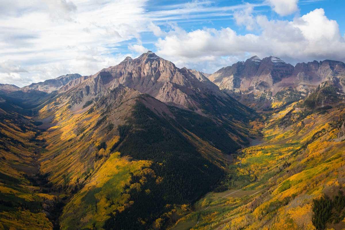 An aerial view of rocky mountain peaks during the fall. At lower elevations the mountains are covered in golden trees, at higher altitudes the mountains are sheer with a bit of snow.