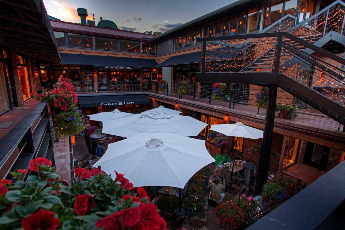 In a courtyard of a burnt-orange building, dining tables are shielded from the sun with white umbrellas.