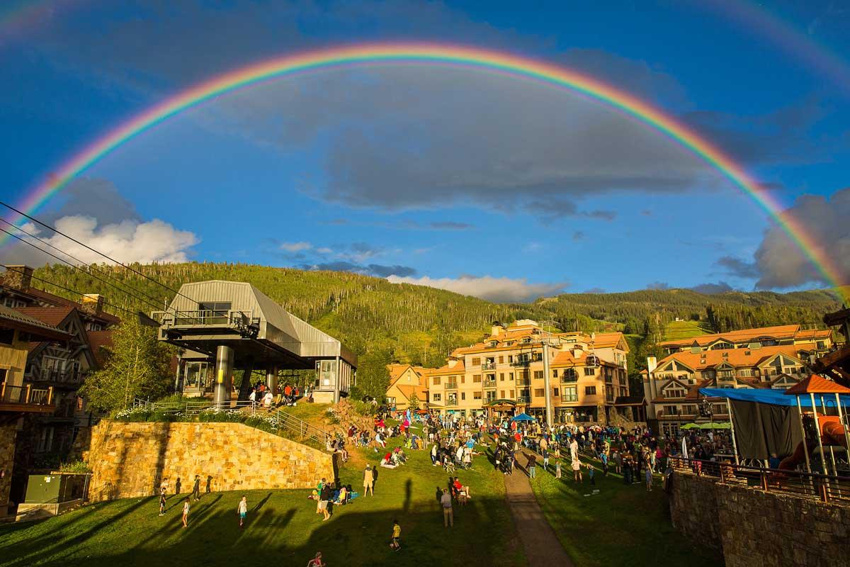 A rainbow over a bustling ski area with a blue sky up above.