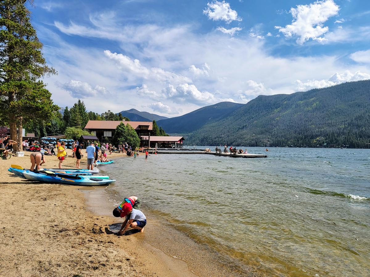 Boats and swimmers mingle on the shores of Grand Lake in Colorado with a wooden dock and forested mountains in the background.