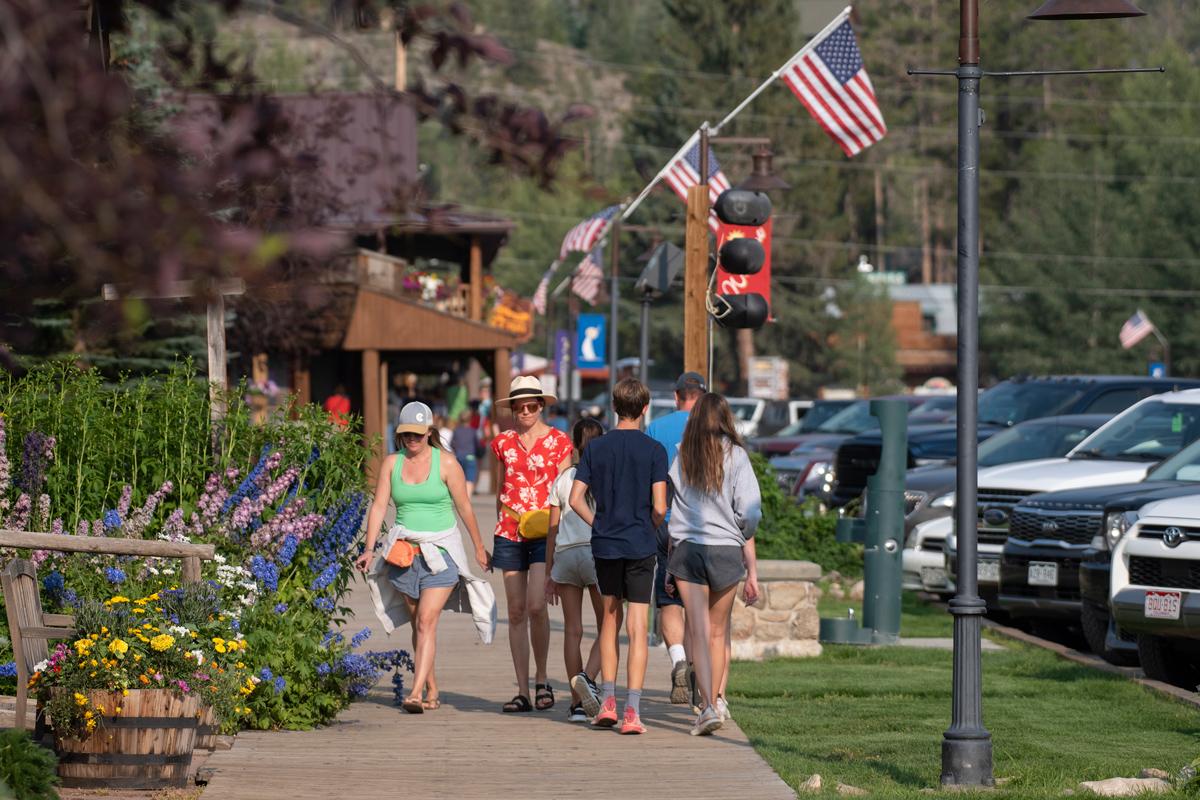 People stroll along the historical boardwalk in Grand Lake, Colorado, with American flags attached to posts overhead and vibrant mini-gardens of flower nearby.