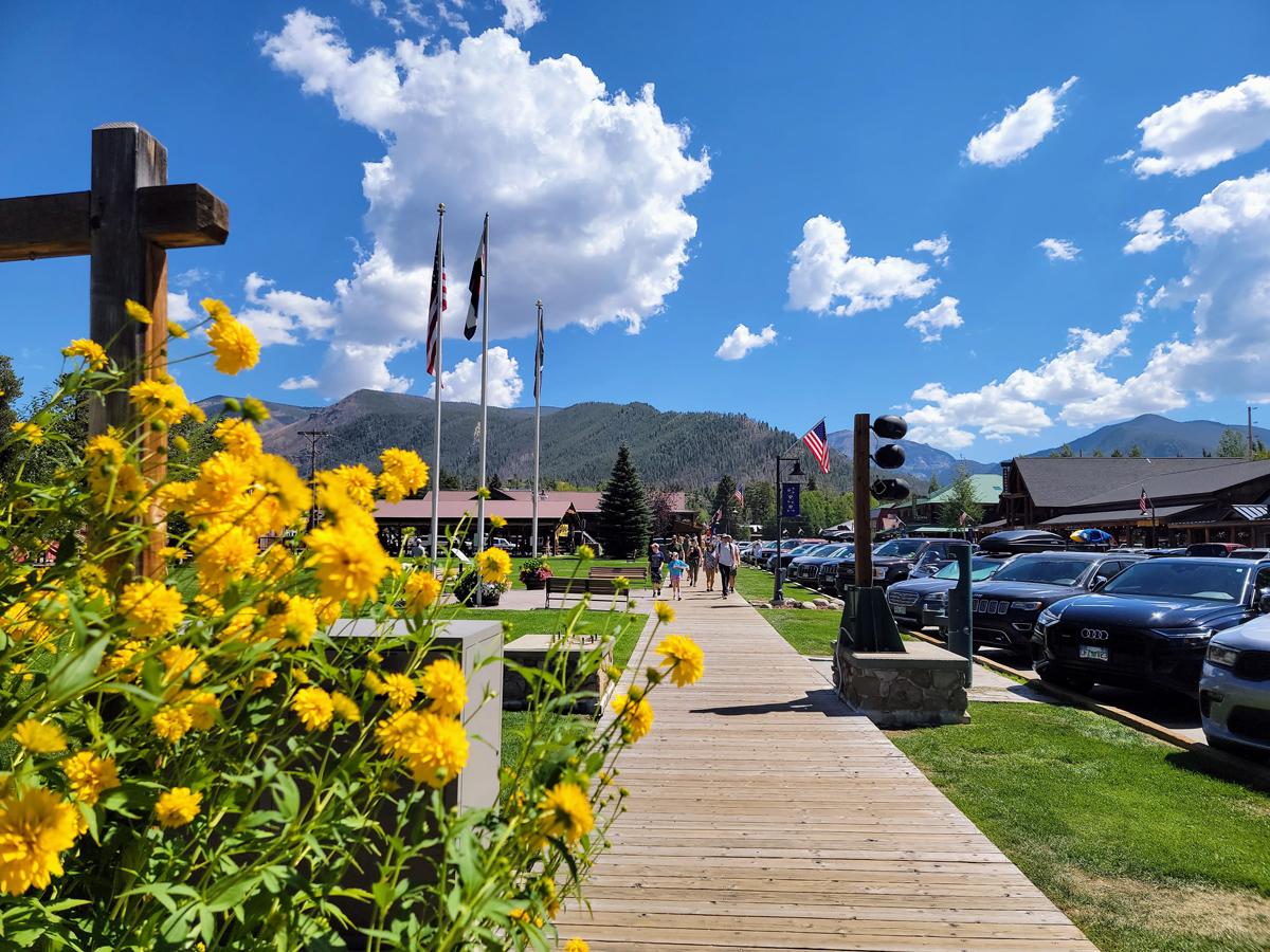 On a blue-sky, summer's day with white clouds, people walk down a historical boardwalk in Grand Lake. Yellow flowers grow in abundance on the left side of the image.