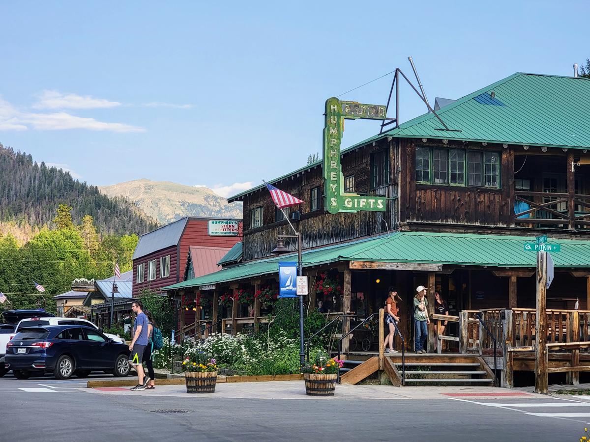 On the corner of Grand Avenue, a two-story wooden building with a green roof sits with people mingling outside. There's a blue-sky with mountains in the background.